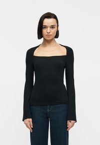 Γυναίκα που φοράει μαύρη μακρυμάνικη ριπ μπλούζα με τετράγωνο ντεκολτέ και φαρδιές ανοίγματα στους ώμους, συνδυασμένη με σκούρο μπλε τζιν, στέκεται μπροστά σε λευκό φόντο.