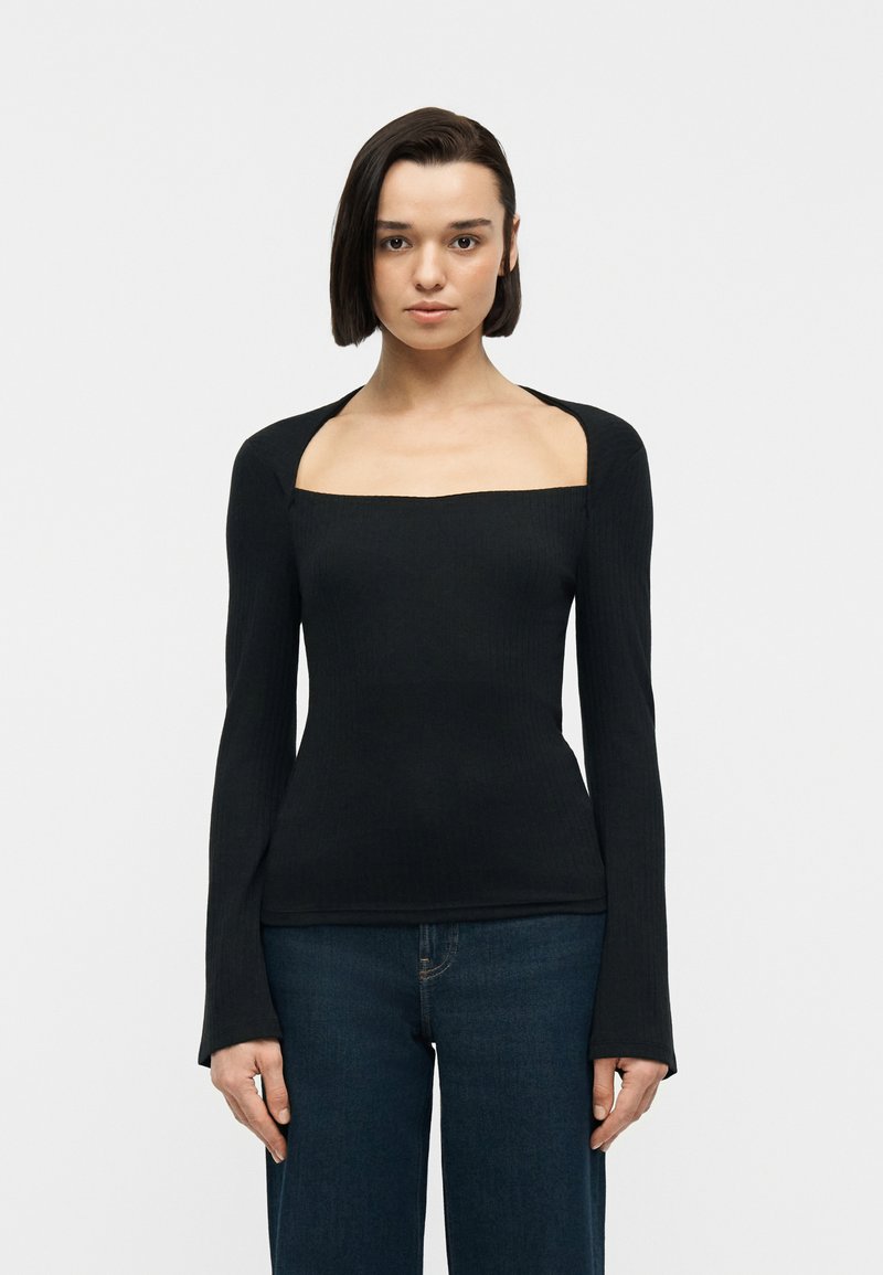 Γυναίκα που φοράει μαύρη μακρυμάνικη ριπ μπλούζα με τετράγωνο ντεκολτέ και φαρδιές ανοίγματα στους ώμους, συνδυασμένη με σκούρο μπλε τζιν, στέκεται μπροστά σε λευκό φόντο.