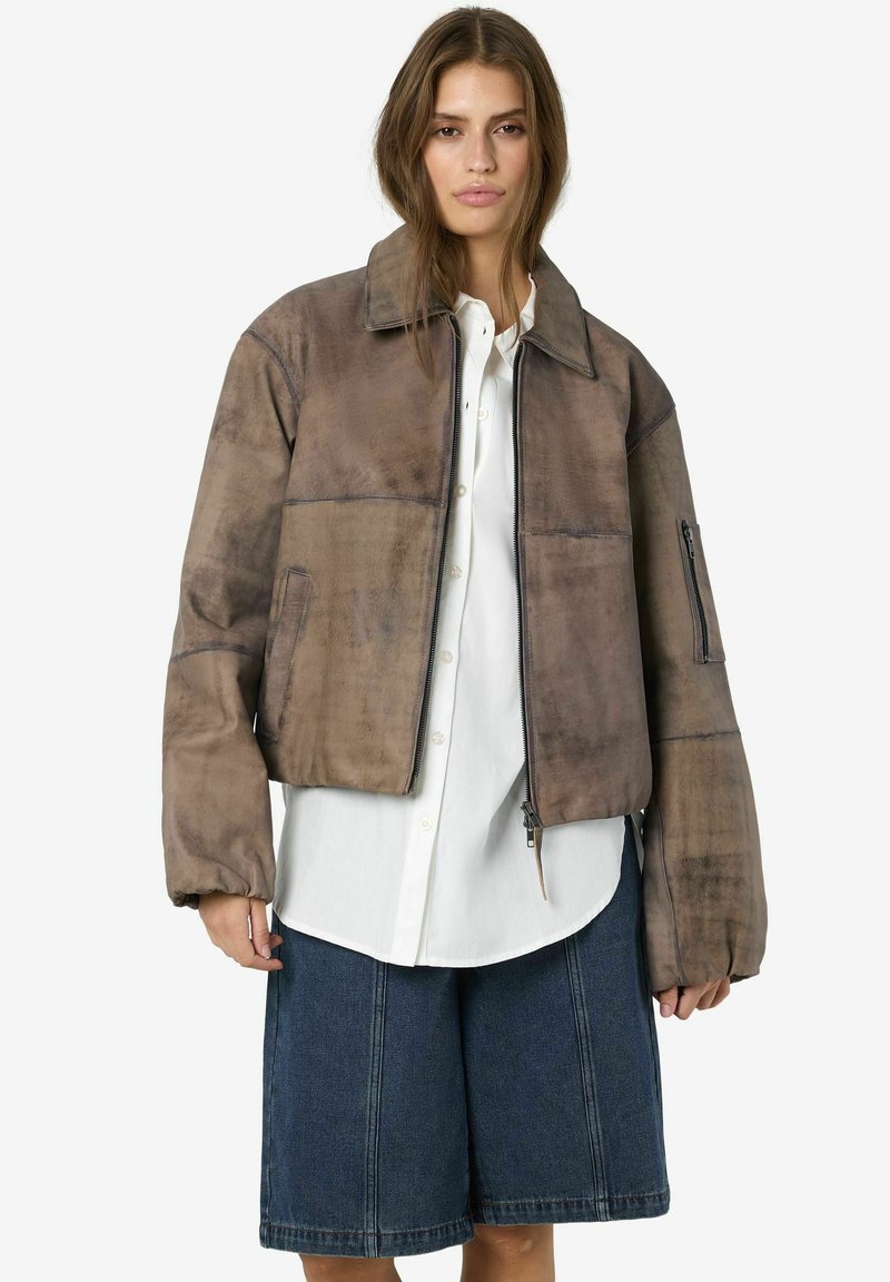 Jeune femme portant une veste en daim marron surdimensionnée, une chemise blanche à boutons et un short en denim longueur genoux sur un fond uni.