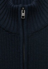 Sininen neulepaita, jossa on teksturoitu kuvio, korkealla ribbikauluksella ja keskellä metallivetoketju, jossa on metallinen vetoketjun vetäjä.