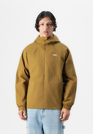 Brun hettegenser med glidelås foran, elastiske mansjetter og en logo på brystet. Bært med lys blå denimbukser.