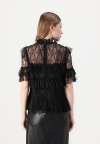Top in pizzo nero con volant a livelli, maniche corte e colletto alto. Chiusura con zip sul retro, design trasparente e tessuto testurizzato.