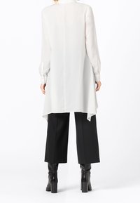 Camicia bianca a maniche lunghe con orlo asimmetrico, realizzata in materiale leggero. Abbinata a pantaloni neri a gamba larga e stivali neri con tacco.