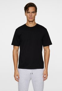 T-shirt en coton noir avec un col rond et des manches courtes, arborant un logo discret sur la poitrine, porté avec un pantalon de jogging gris clair.