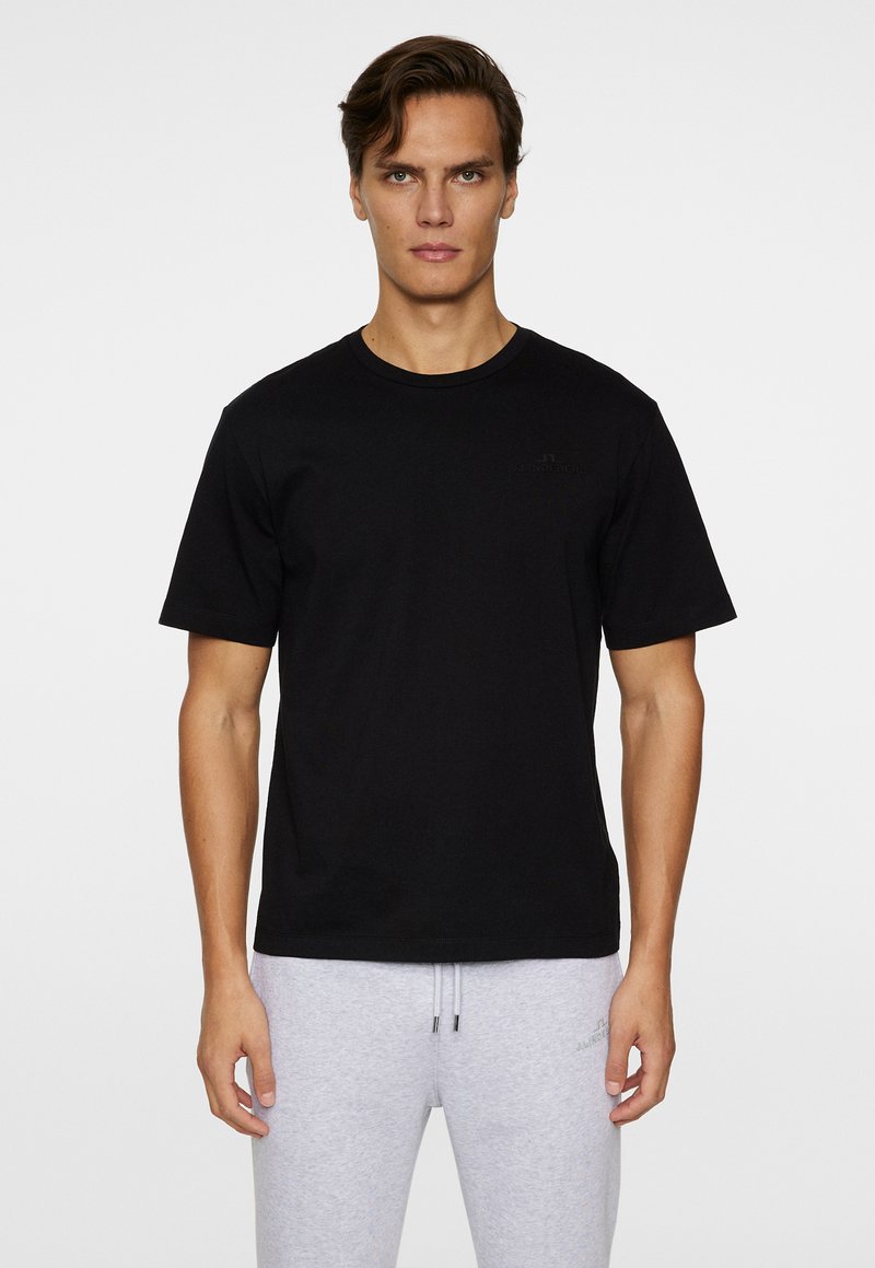 T-shirt en coton noir avec un col rond et des manches courtes, arborant un logo discret sur la poitrine, porté avec un pantalon de jogging gris clair.