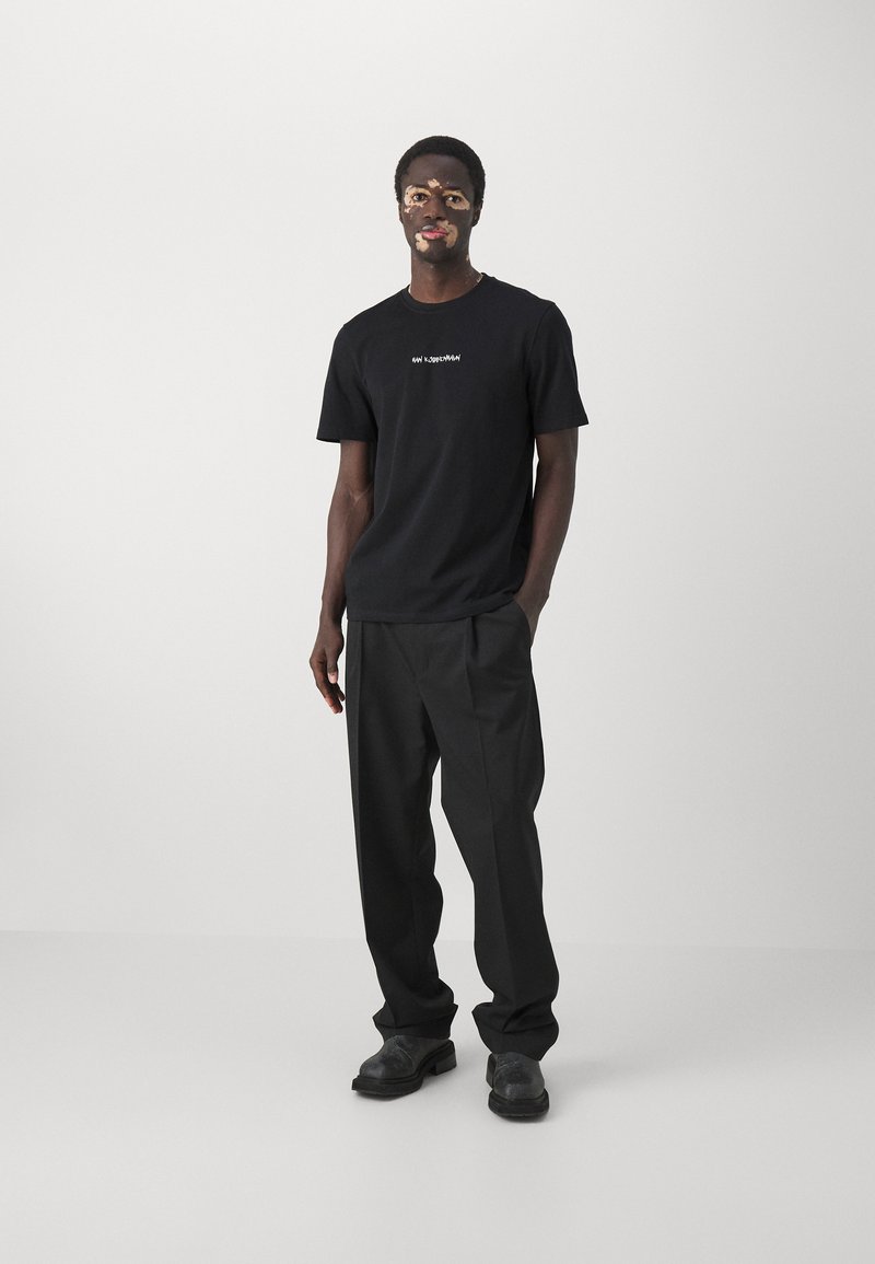 Tricou negru din bumbac, cu mâneci scurte și detalii albe, purtat cu pantaloni negri largi și încălțăminte neagră. Textură netedă.