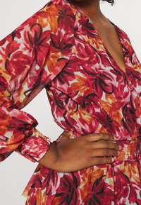 Blusa floral con mangas abullonadas, escote en V profundo y frunces en la cintura. Los colores incluyen rojo, naranja y negro sobre un fondo claro.
