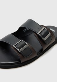 Sandale à glissiére en cuir noir et marron avec deux sangles ajustables et boucles en argent, semelle plate, texture lisse et branding subtil.