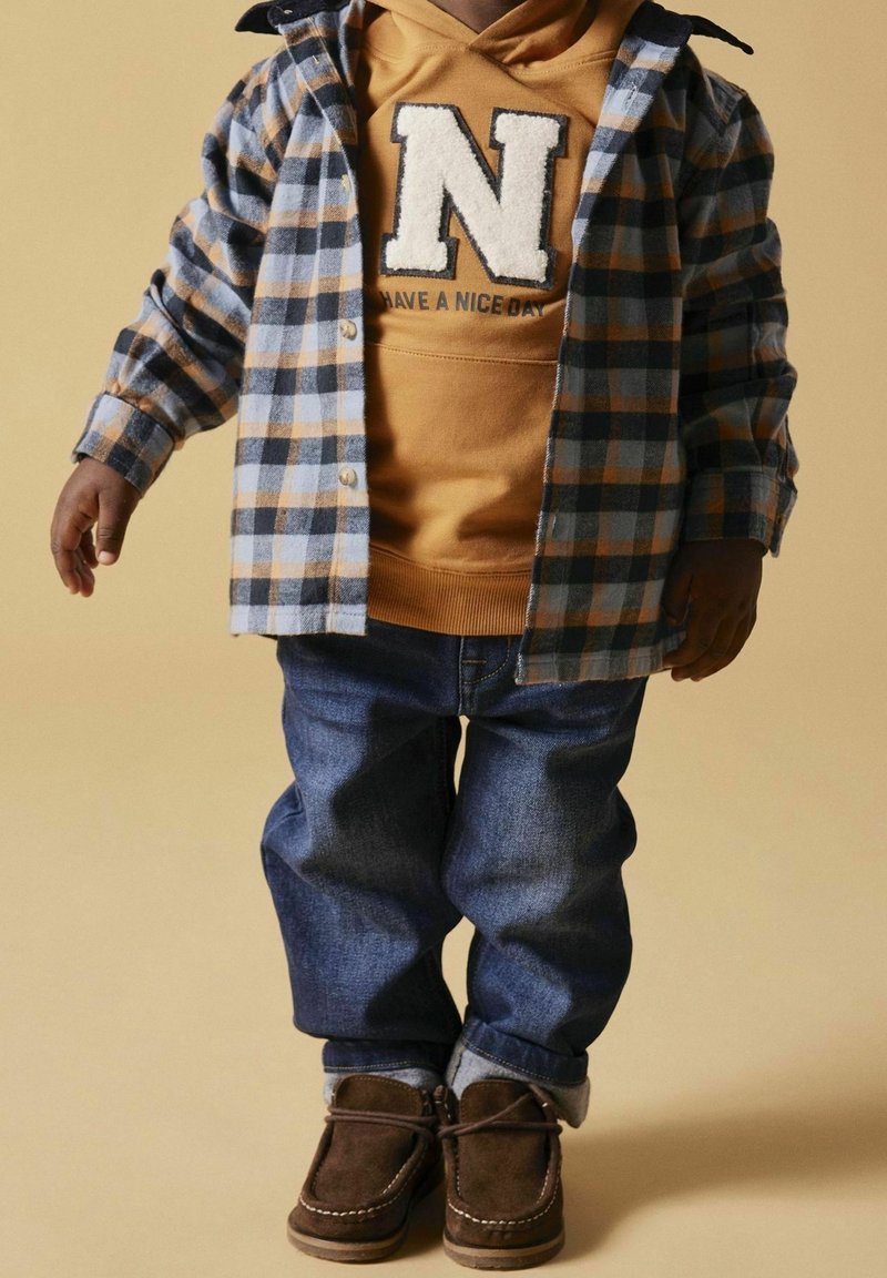 Bambino che indossa una camicia a quadri blu e grigia sopra una felpa giallo senape con la lettera "N" e la scritta "Have a nice day", jeans blu e mocassini marroni.