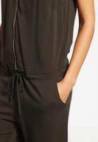 Olivegrüner Jumpsuit mit einem Reißverschluss vorn, verstellbarem Taillengürtel und seitlichen Taschen. Aus glattem, leichtem Stoff gefertigt.