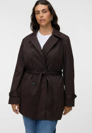Femme portant un trench-coat marron foncé ceinturé par-dessus une chemise blanche et un jean bleu, debout devant un fond clair uni.