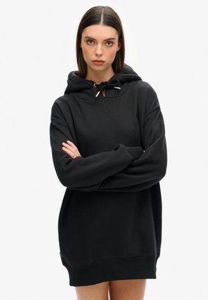 Junge Frau mit glattem braunem Haar trägt übergroßes schwarzes Hoodie-Kleid, die Arme verschränkt, steht vor einfachem hellem Hintergrund.