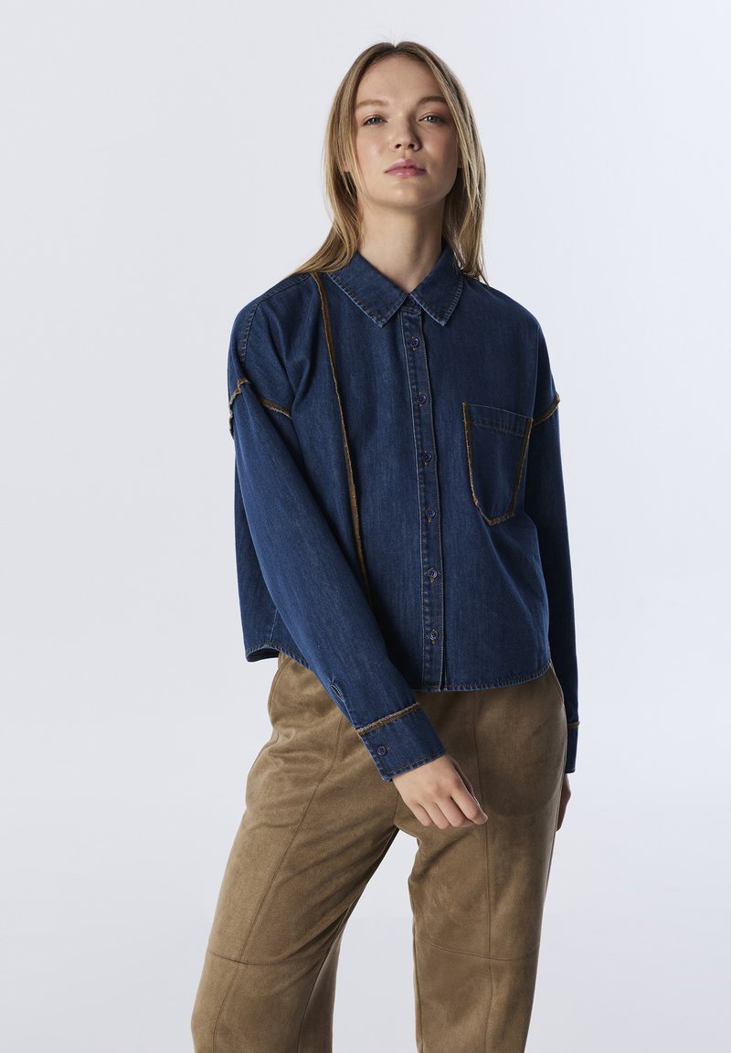 Blauwe denim shirt met een knoopsluiting aan de voorkant, kraag, borstzak en contrasterende gele stiksels. Gecombineerd met beige suède broeken.
