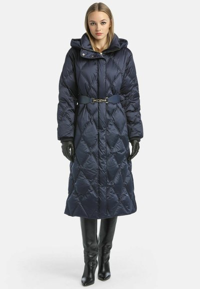 Geacă puffer matlasată de culoare bleumarin, cu glugă, talie cu curea și model în diamant; purtată cu mănuși negre și cizme până la genunchi.
