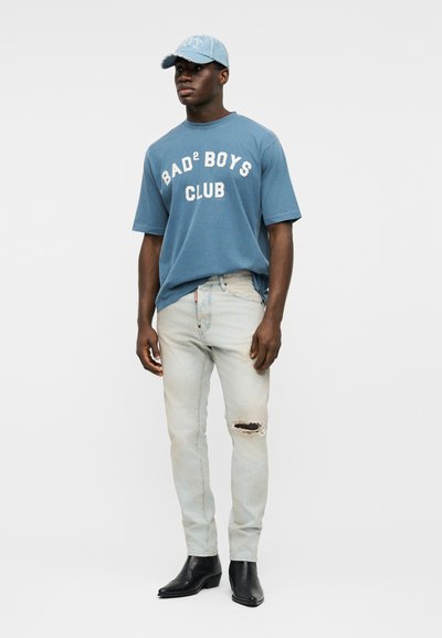 Jeune homme portant un t-shirt bleu "BAD² BOYS CLUB", un jean clair déchiré, des bottes noires et une casquette bleue assortie, debout devant un fond uni.