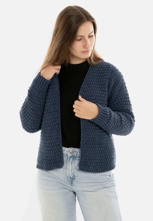 Donkerblauwe gebreide cardigan met een textuurpatroon, open voorkant en geribbelde manchetten, gecombineerd met een zwarte top en lichtblauwe spijkerbroek.