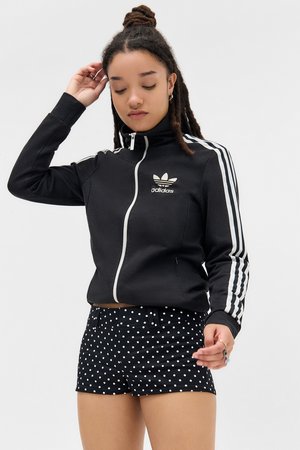 Femme avec des dreadlocks portant une veste zippée noire Adidas avec des rayures blanches et un short à pois noirs, ajustant ses cheveux d'une main.