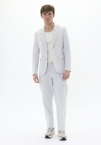 Veste de costume gris clair à rayures fines et pantalon assorti, associés à un débardeur beige et des baskets de sport neutres sur un fond uni.