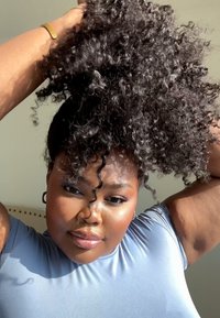 Kihara musta hiukset kiinni, yllään vaaleansininen lyhythihainen paita. Pehmeä iho lämpimässä valaistuksessa, esittäen määriteltyjä kiharoita ja hienoista kiiltoa.