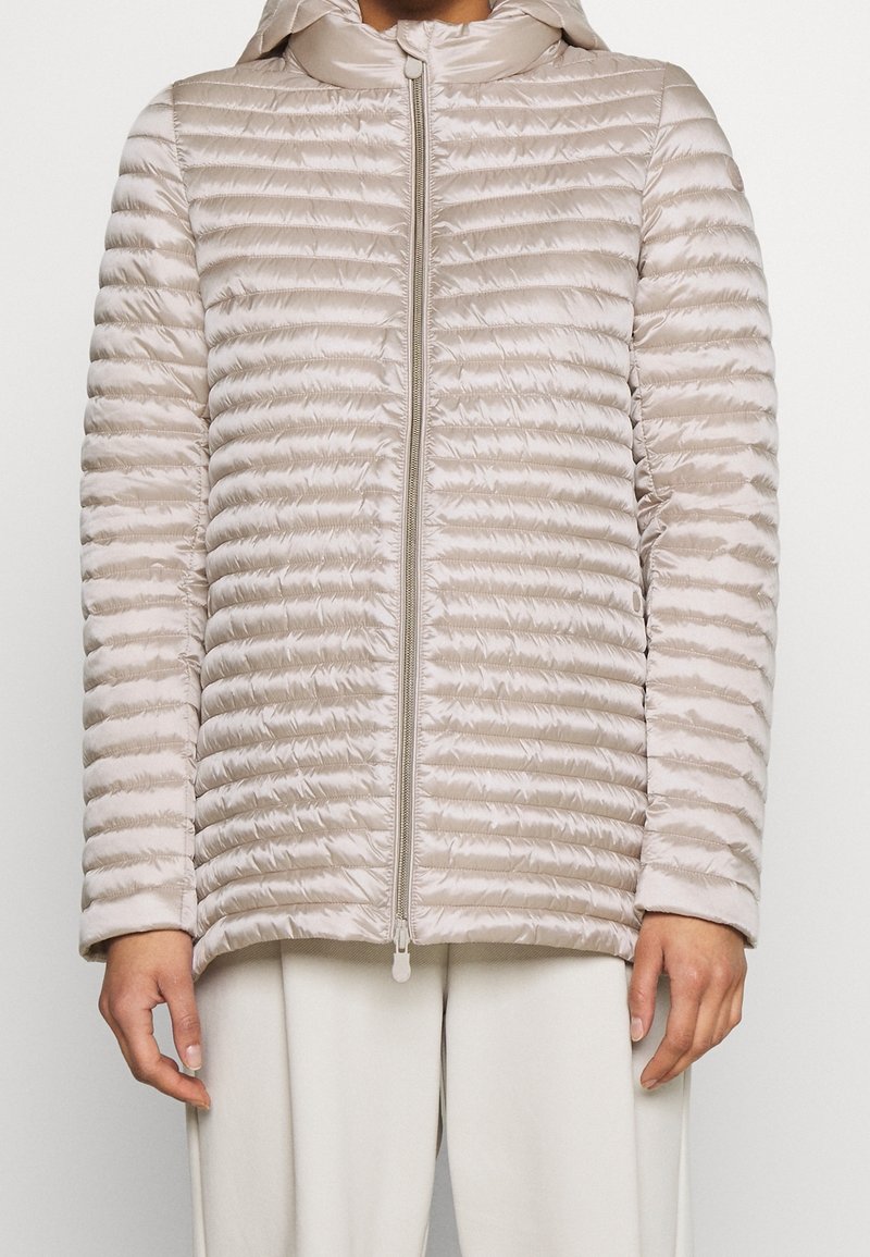 Beżowa pikowana kurtka z kapturem, z przeszyciami, przednim zamkiem błyskawicznym i gładką, błyszczącą fakturą. Noszona na jasnych spodniach.