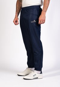 Marine windbreaker broek met een elastische tailleband, tapered fit en zijzakken met rits. Logo gedrukt op de linker bovenbeen. Witte sneakers.