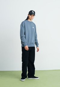 Felpa grigia con scollatura rotonda e piccolo logo, abbinata a pantaloni neri larghi e sneakers nere. Outfit semplice e casual.