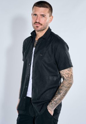 Mann mit kurzen Haaren und Tattoos, der ein schwarzes kurzärmeliges Hemd über einem weißen T-Shirt trägt und mit einer Hand in der Hosentasche steht.