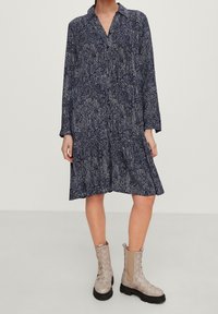 Marinblå klänning med långa ärmar, krag neckline och vit mönstrad design; kombinerad med ljusbeige ankelskor med vävd textur.