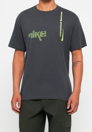 Mand iført mørkegrå Nike T-shirt med neongrøn "nike" tekst og lodret "International Sportswear" tekst med swoosh-logo på brystet.