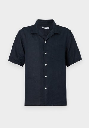 Muž s krátkými blond vlasy, který má na sobě černou košili s krátkým rukávem na knoflíky, bílý nátělník, béžové široké kalhoty a tmavé sandály.