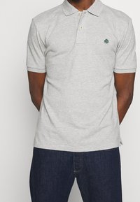 Vaaleanharmaa puuvillapolo, jossa on kaulus, kaksi kultanappia ja pieni vihreä puulogo. Lyhyet hihat ja suora malli.