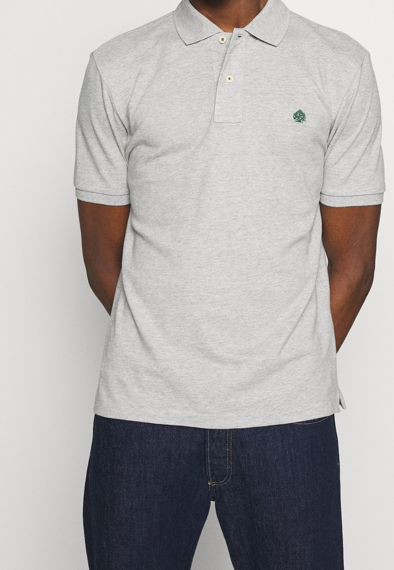 Vaaleanharmaa puuvillapolo, jossa on kaulus, kaksi kultanappia ja pieni vihreä puulogo. Lyhyet hihat ja suora malli.