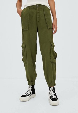 Persoon draagt olijfgroene cargobroek met elastische boorden en zwarte hoge sneakers met witte veters, handen in de zakken.