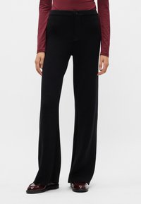 Mustad laiad püksid siledast materjalist, elastse vöökoha, nööbiga ja lõdva lõikega. Ühendatud tumedate kingade ja kitsa Ülaosaga.