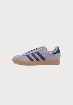 Ανοιχτό γκρι σουέτ αθλητικό παπούτσι Adidas Gazelle με ριγέ σε ναυτικό μπλε και λαστιχένια σόλα, εμφανίζεται από την πλευρά σε απλό φόντο.