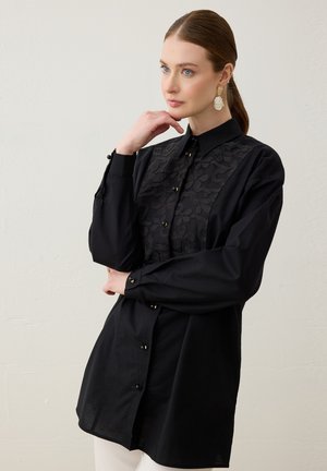 Femme à la peau claire et aux cheveux bruns attachés, portant une chemise noire à manches longues avec broderie florale et des boucles d'oreilles en or, posant avec la main près du visage.