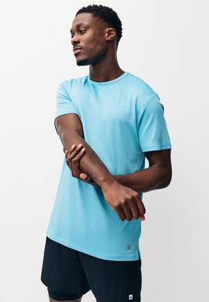 Jeune homme en chemise bleu clair à manches courtes et short noir étirant son bras gauche à travers sa poitrine devant un fond uni.