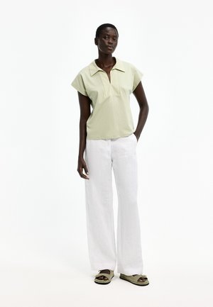 Frau trägt ein hellgrünes kurzärmeliges Oberteil mit Kragen, weiße Hosen mit weitem Bein und passende grüne Sandalen, steht vor einem weißen Hintergrund.