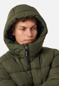Oliwkowa kurtka puchowa z dużym kapturem, teksturowanym pikowaniem i zamek błyskawiczny z przodu. Posiada regulowane sznurki do dopasowania.