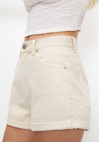 Shorts en denim couleur crème au design ajusté, avec un ourlet retroussé et des poches avant. Comprend un bouton en métal et des détails de couture subtils.
