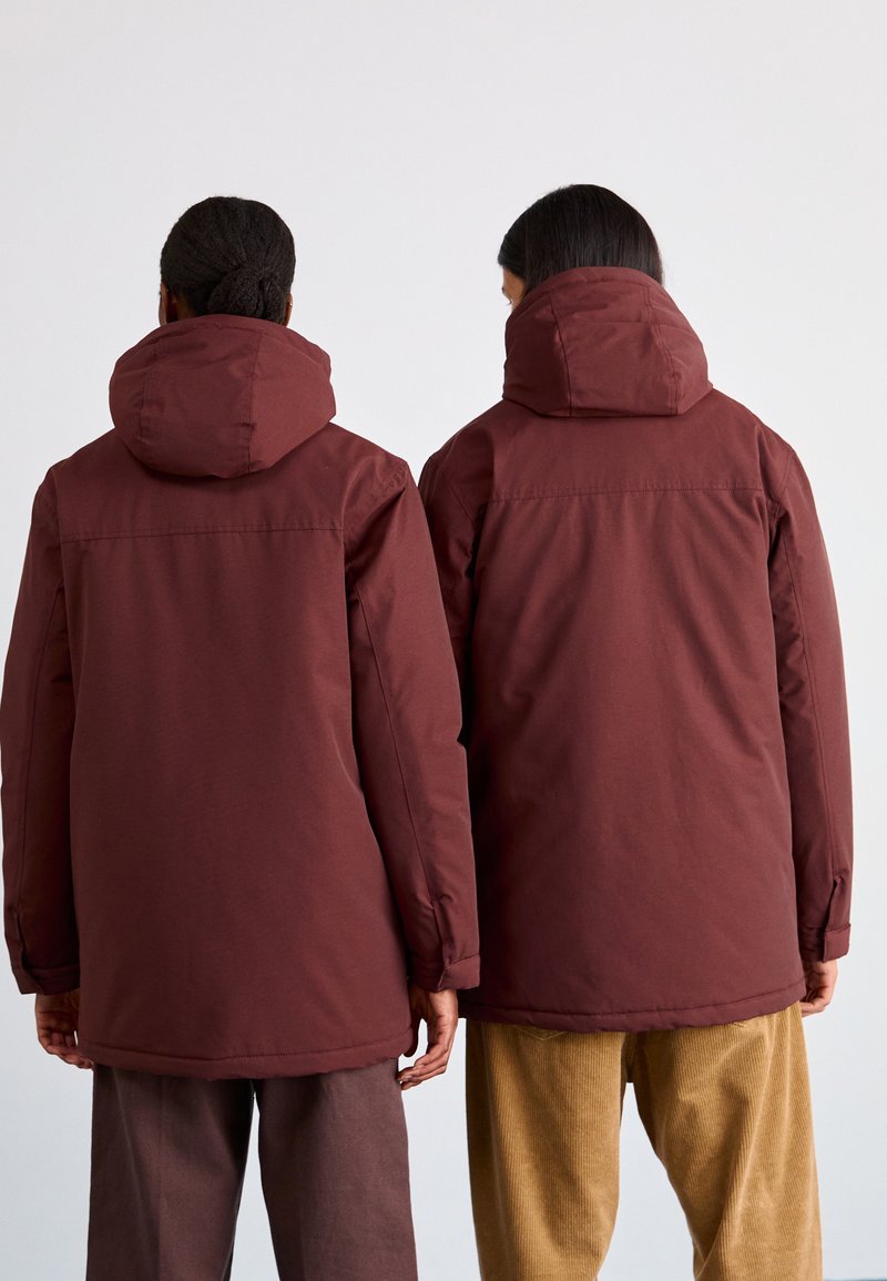 Chaquetas aislantes de color burdeos con capuchas, que presentan una textura lisa y un corte recto, usadas por dos personas vistas desde atrás.