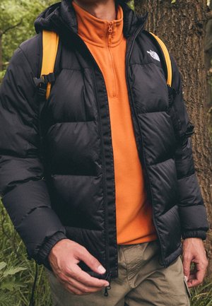 Mann trägt einen orangefarbenen Reißverschluss-Pullover, eine schwarze Steppjacke und gelbe Rucksackriemen, steht im Freien in der Nähe eines Baumes.