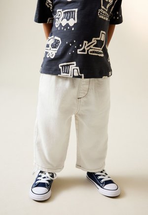 Barn iført hvide bukser, mørkeblå sneakers og en sort t-shirt med hvide tryk af byggepladskøretøjer.