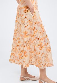 Γυναίκα που φοράει μπεζ midi φούστα με πορτοκαλί floral μοτίβο, στέκεται με το ένα χέρι στην τσέπη, σε συνδυασμό με ανοιχτό μπεζ slingback flats.