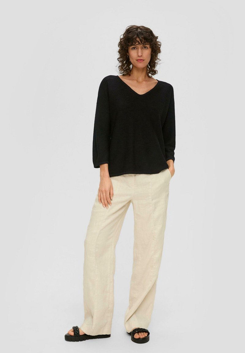 Schwarzer V-Ausschnitt-Pullover mit langen Ärmeln, kombiniert mit lockeren beigen Leinenhosen und schwarzen Plateau-Sandalen. Glatte Textur, entspannter Schnitt.