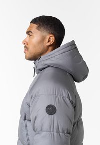Chaqueta de plumas gris con capucha, tejido texturizado y un parche de logotipo circular en el hombro izquierdo. Costuras de calidad visibles.