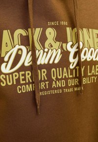 Brun sweatshirt med en tekstureret stof, der har hvid og gul grafisk tekst, der siger "Jack & Jones Denim Goods" og "Superior Quality Label."