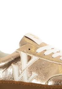 Metalen gouden sneaker met witte accenten, een textuur oppervlak en een opvallend logo aan de zijkant. Witte veters en een ronde neusontwerp.