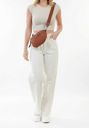 Braune Leder-Umhängetasche mit geschwungener Form, Reißverschluss und verstellbarem Riemen, kombiniert mit weißen Hosen und weißen Turnschuhen.