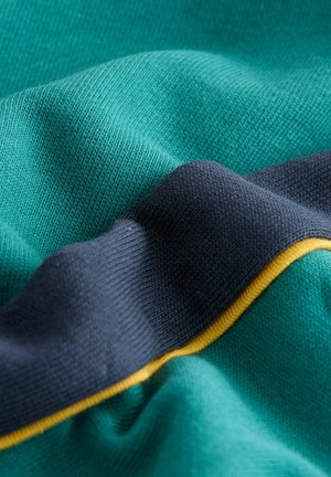 Nahaufnahme eines türkisfarbenen Strickstoffs mit einem marineblauen Streifen, der von einer dünnen gelben Linie umrandet ist, wobei die Textur und Falten sichtbar sind.